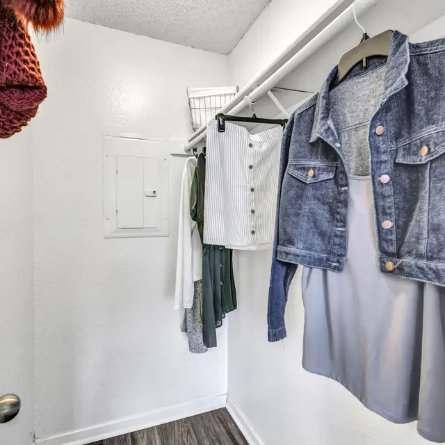 A small closet with hanging clothes including a denim jacket, shirt, and various garments against a white wall.