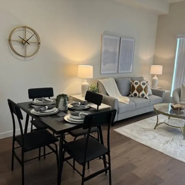 A stylish living room and dining area featuring a dining table set for four, modern furniture, and decorative items. A light-colored sofa with decorative pillows, a glass coffee table, and plants are visible in the space. A large clock is mounted on the wall, and large windows provide natural light.