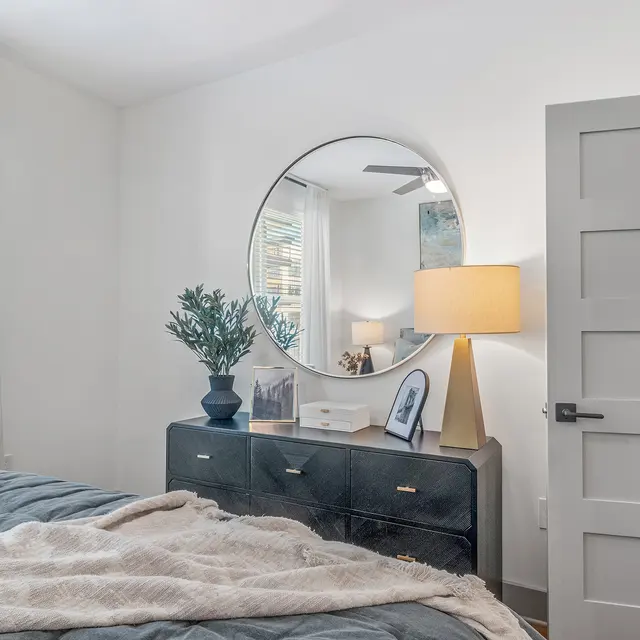 Stylish Modern Bedroom A modern bedroom featuring a bed with a blue comforter, a round mirror above a dark dresser, and a lamp. The room has light-colored walls and curtains, adding a bright and airy feel.