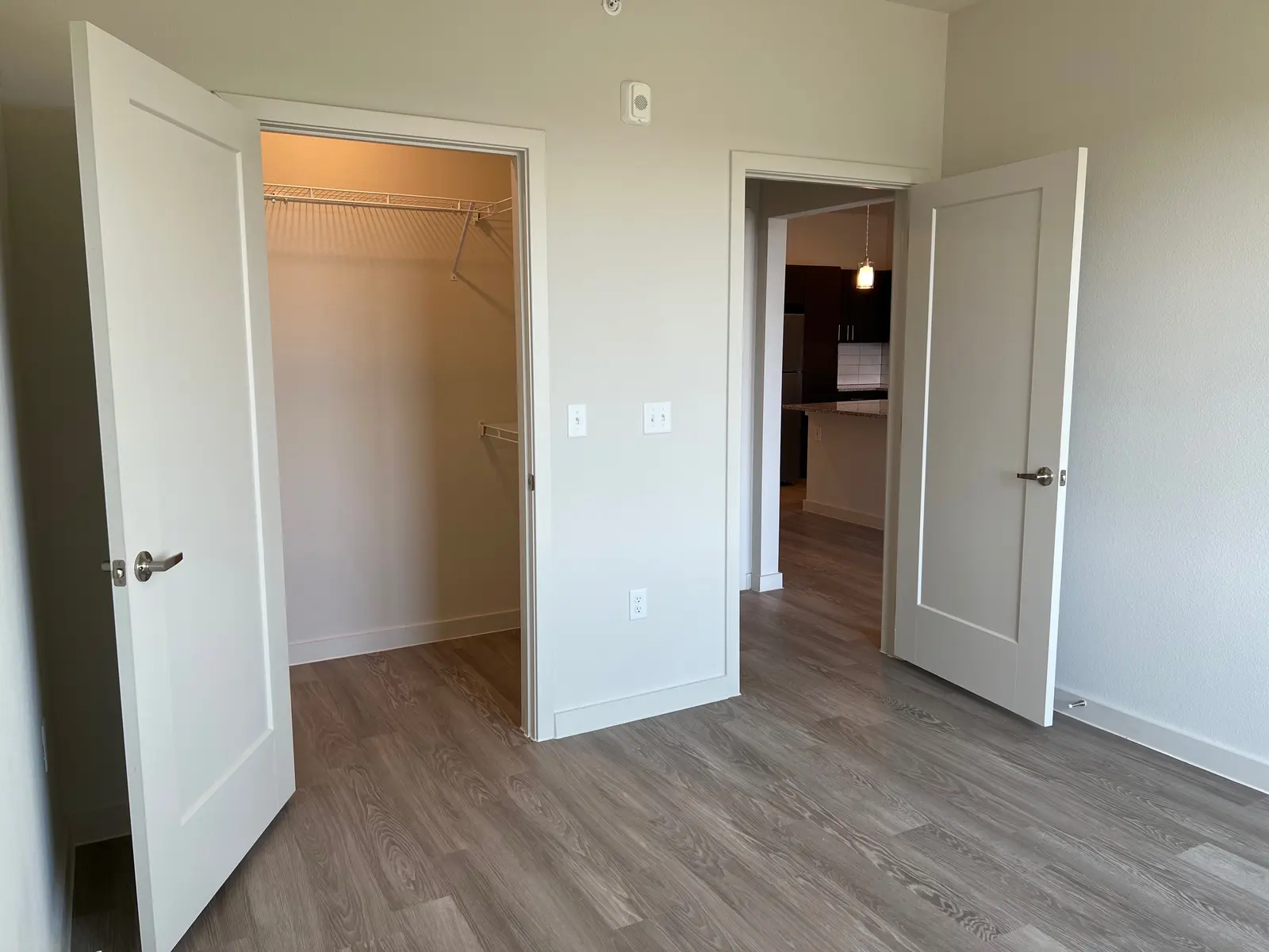An empty room featuring two open doors; one leads to a closet and the other to a kitchen area in the background. The floor is covered with light wood-like flooring and the walls are painted light gray.