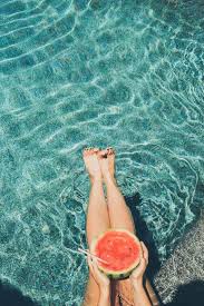 A person's legs are submerged in clear blue water, holding a large slice of watermelon.
