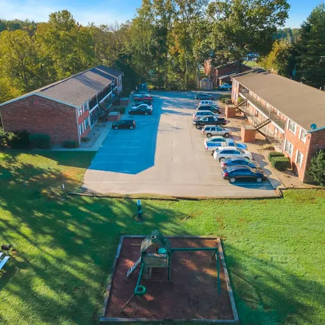 Aerial view of a brick apartment complex surrounded by trees and greenery. There is a parking area in front of the buildings and a playground with equipment in the foreground.