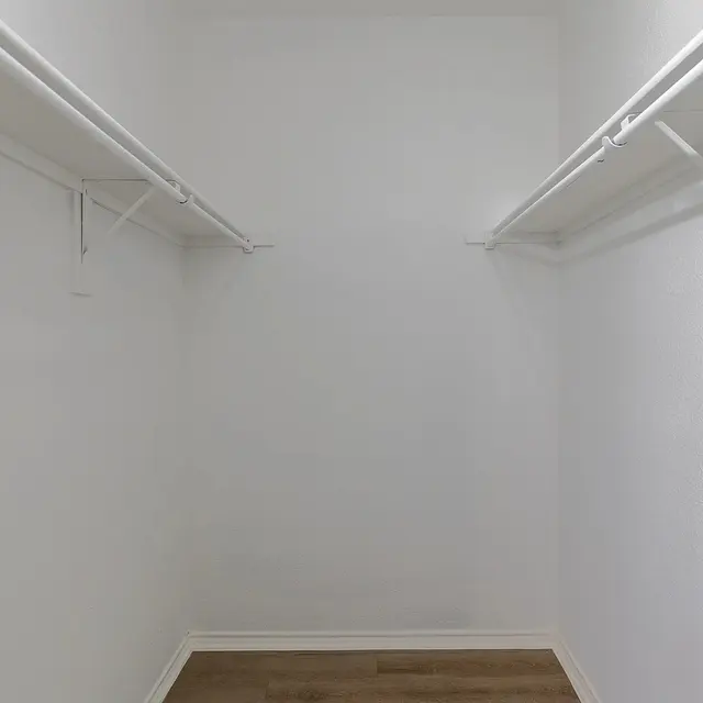 An empty closet with white walls and wooden flooring, featuring two hanging rods on opposite walls.
