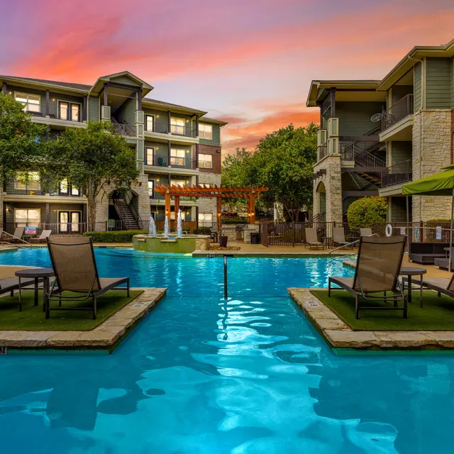 A serene outdoor apartment pool area at sunset, featuring lounge chairs, green trees, and buildings in the background.
