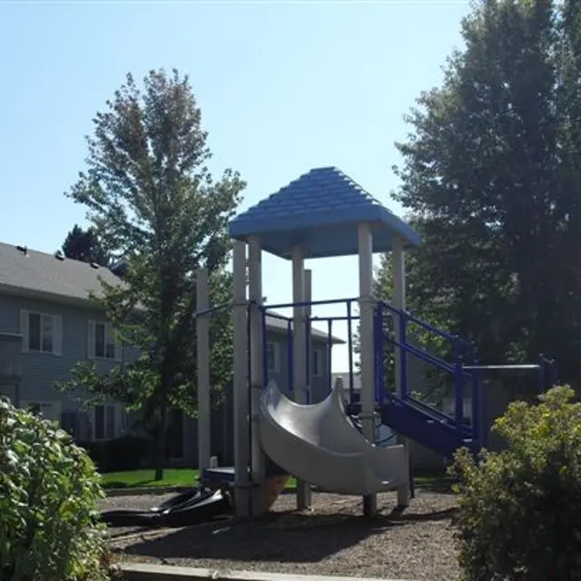 Playground with Slide A playground featuring a blue slide and structure surrounded by trees, with apartment buildings in the background.