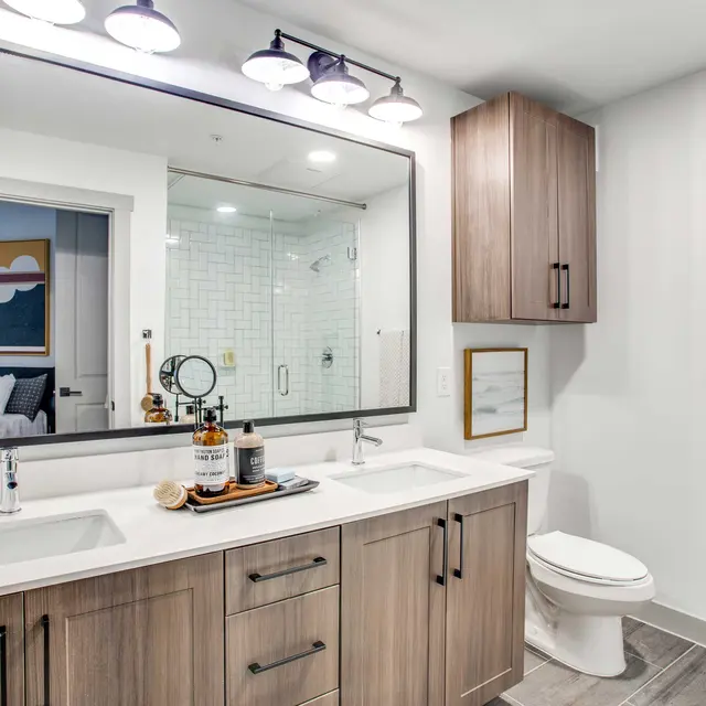 A modern bathroom featuring a double sink vanity with wooden cabinets, a large mirror with light fixtures above, a glass shower stall in the background, and decorative items on the countertop.