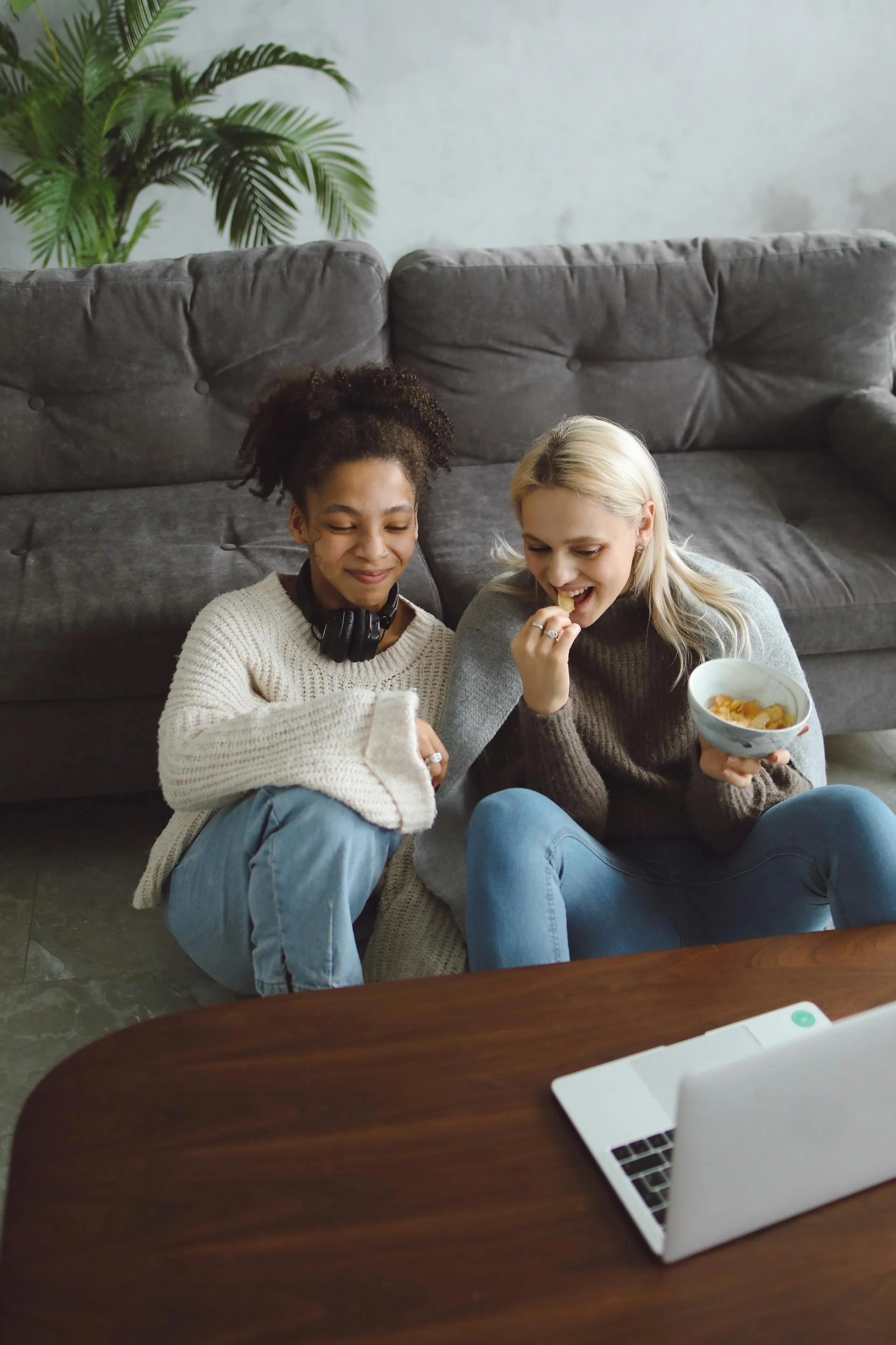Cozy Movie Night with Friends Two friends sitting on the floor in cozy clothing, enjoying snacks while watching something on a laptop. They are surrounded by a comfortable living room setting with a large sofa and indoor plants.