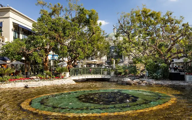 A scenic view of a pond surrounded by lush greenery and trees, with a small bridge crossing over it. The setting features storefronts and a dining area in the background, creating an inviting atmosphere.