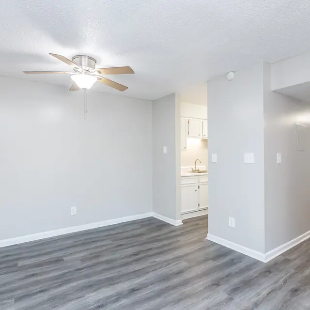 Bright Living Room with Kitchen Access A bright and spacious living room featuring light gray walls and wooden flooring. A ceiling fan is installed, and a pathway leads to a kitchen area in the background.