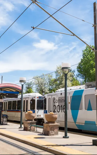 A light rail train parked at a station under a blue sky with scattered clouds. The platform features benches, lamp posts, and signage. Surrounding trees are visible in the background.