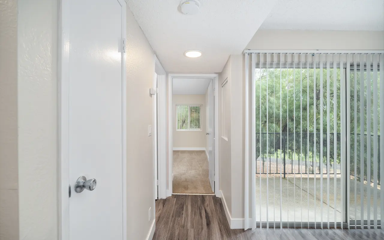 View of a hallway in an apartment with doors on either side and a sliding glass door at the end leading outside.