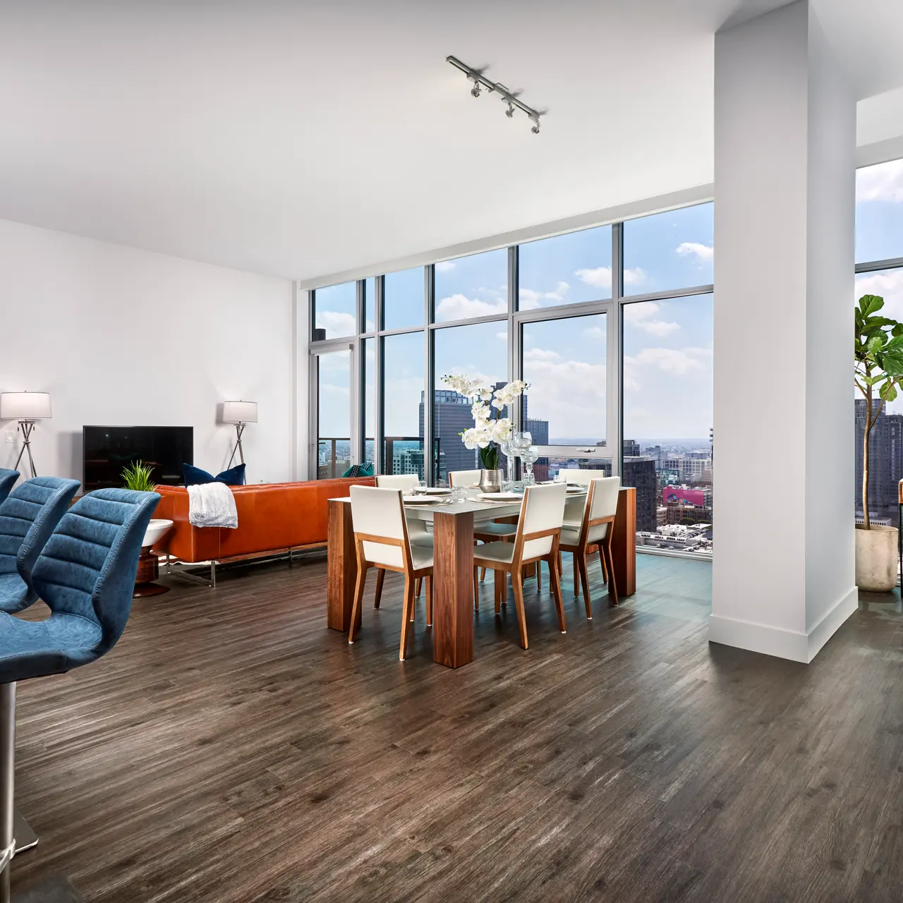 A contemporary open-concept living space featuring a dining area and kitchen. Large windows offer a view of the city skyline, with stylish furniture and decor.