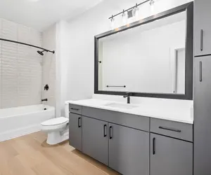 A modern bathroom featuring a gray vanity with white countertops, a large mirror above the sink, and a shower/tub combo with textured tile. The floor is light-colored wood, and the overall decor is minimalist and contemporary.