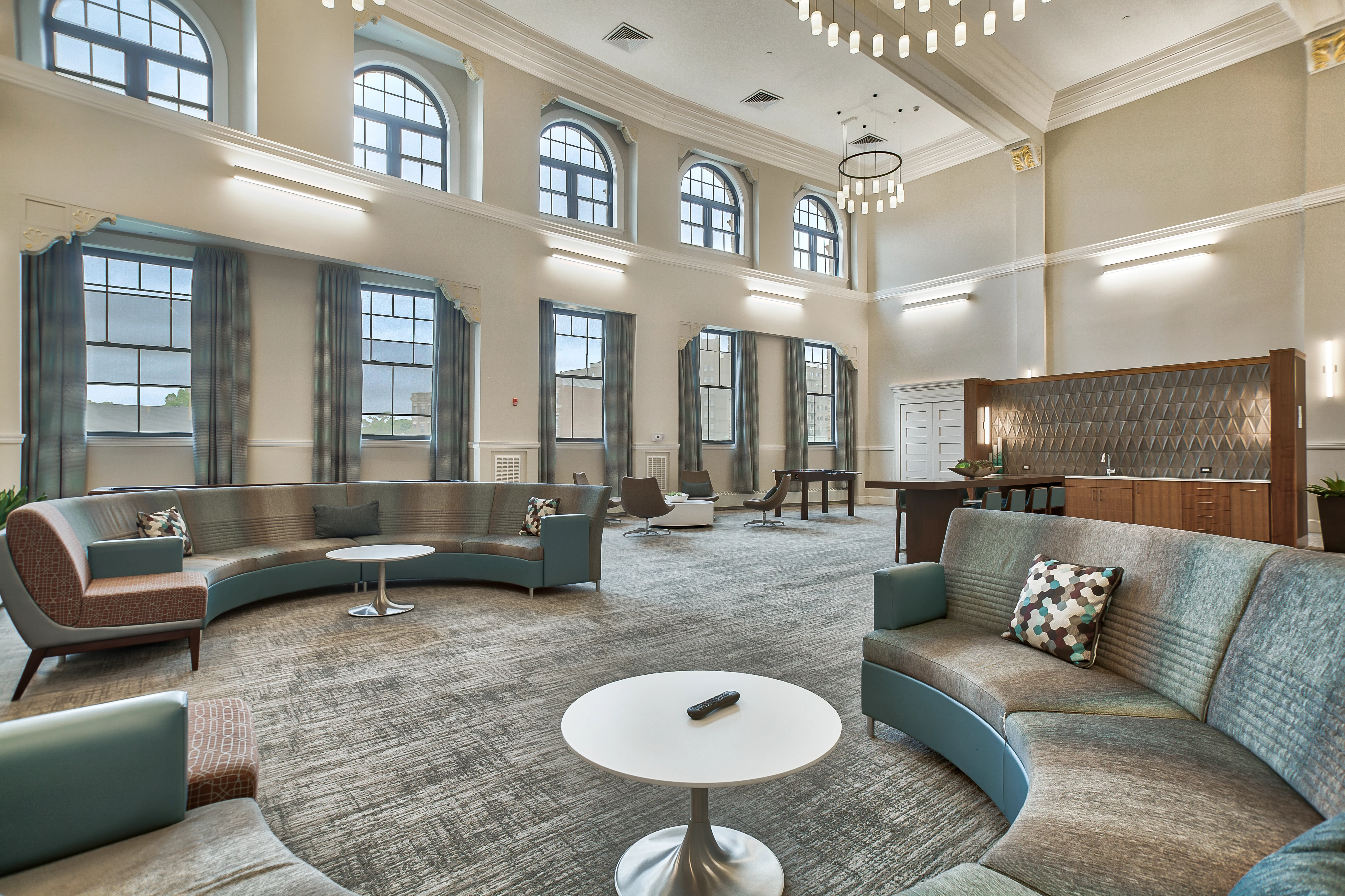 A spacious and modern lobby area featuring curved seating, a round coffee table, large windows, and decorative lighting fixtures.