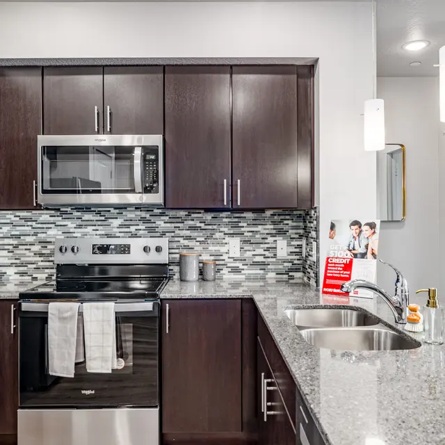 Contemporary Kitchen Design A contemporary kitchen featuring dark wood cabinetry, a stainless steel stove and microwave, and a gray granite countertop with a dual sink. Mosaic tile backsplash adds texture, and attached is a view into a softly lit, stylish living area.