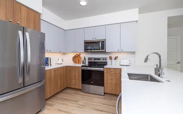A contemporary kitchen featuring stainless steel appliances, wooden cabinetry, and a white countertop. The layout is open with a stainless steel refrigerator, an oven, and a sink with a modern faucet.