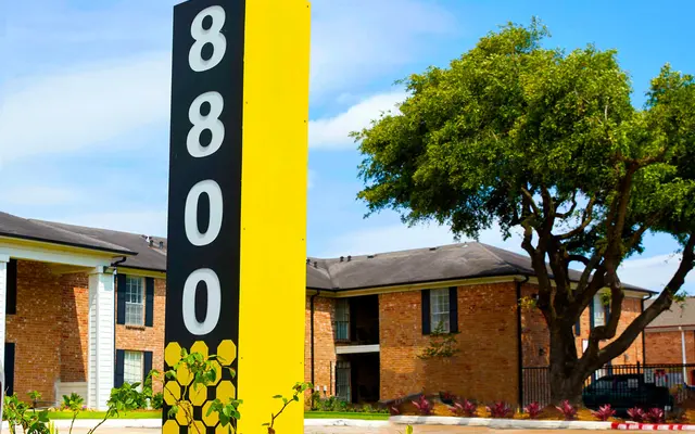 A tall sign displaying the number '8800' in black and yellow, situated in front of an apartment complex with brick exteriors and green foliage in the background.