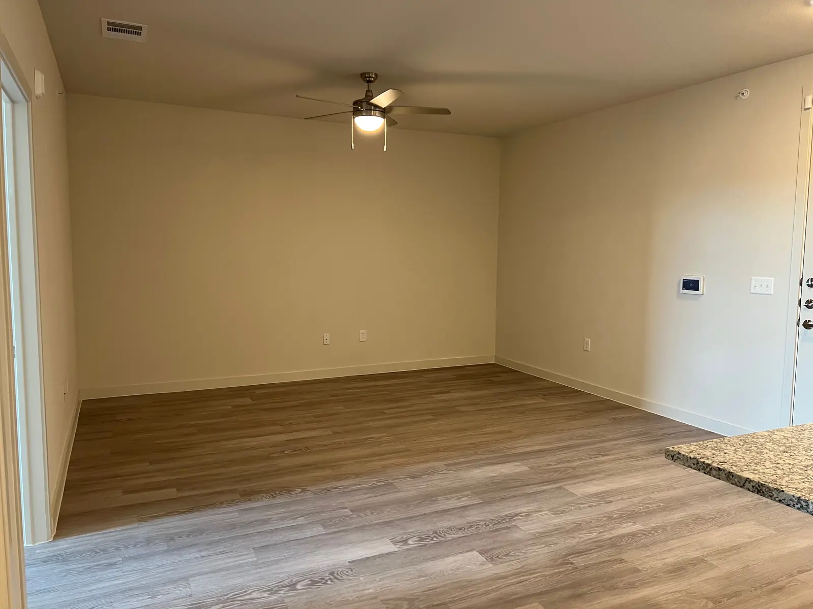 An empty living room with light wood flooring, a ceiling fan, and beige walls. A granite countertop is partially visible on the right side.