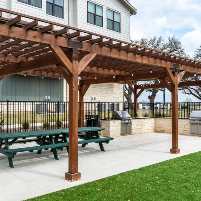 Outdoor patio area with wooden pergola, picnic table, and two grills surrounded by a green lawn and a fence.