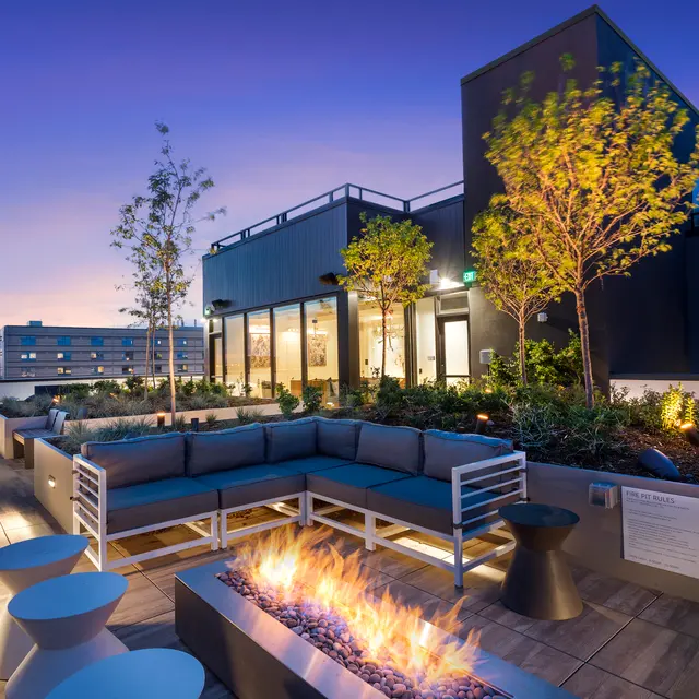 Rooftop lounge with modern seating, greenery, and a fire pit, illuminated against a sunset sky.