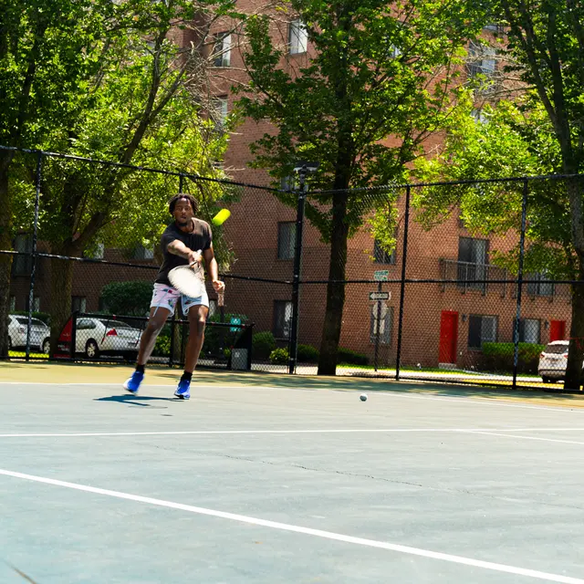 A tennis player in motion about to hit a ball on a sunny court surrounded by trees.