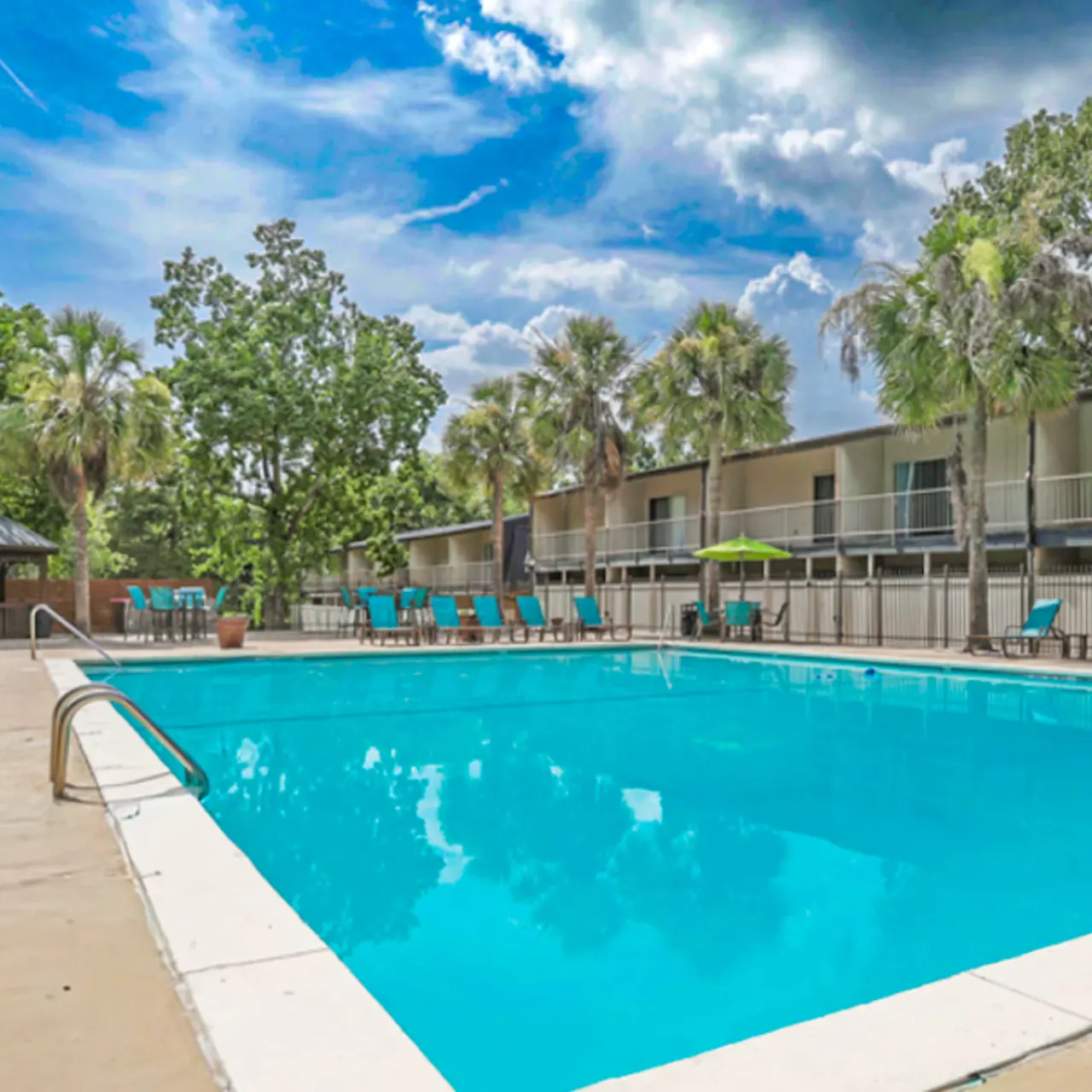 A spacious swimming pool surrounded by lounge chairs and palm trees, with an apartment building in the background under a partly cloudy sky.