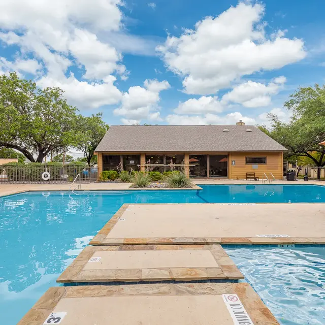 A clear blue swimming pool with a diving board, surrounded by trees and a building. There are tables with umbrellas beside the pool, under a bright blue sky with white clouds.
