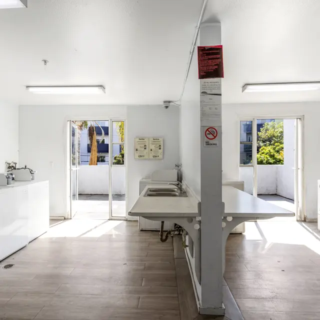 Interior of a bright and clean laundry room featuring several washing machines and dryers. Two doors are visible leading to a bright outdoor area.
