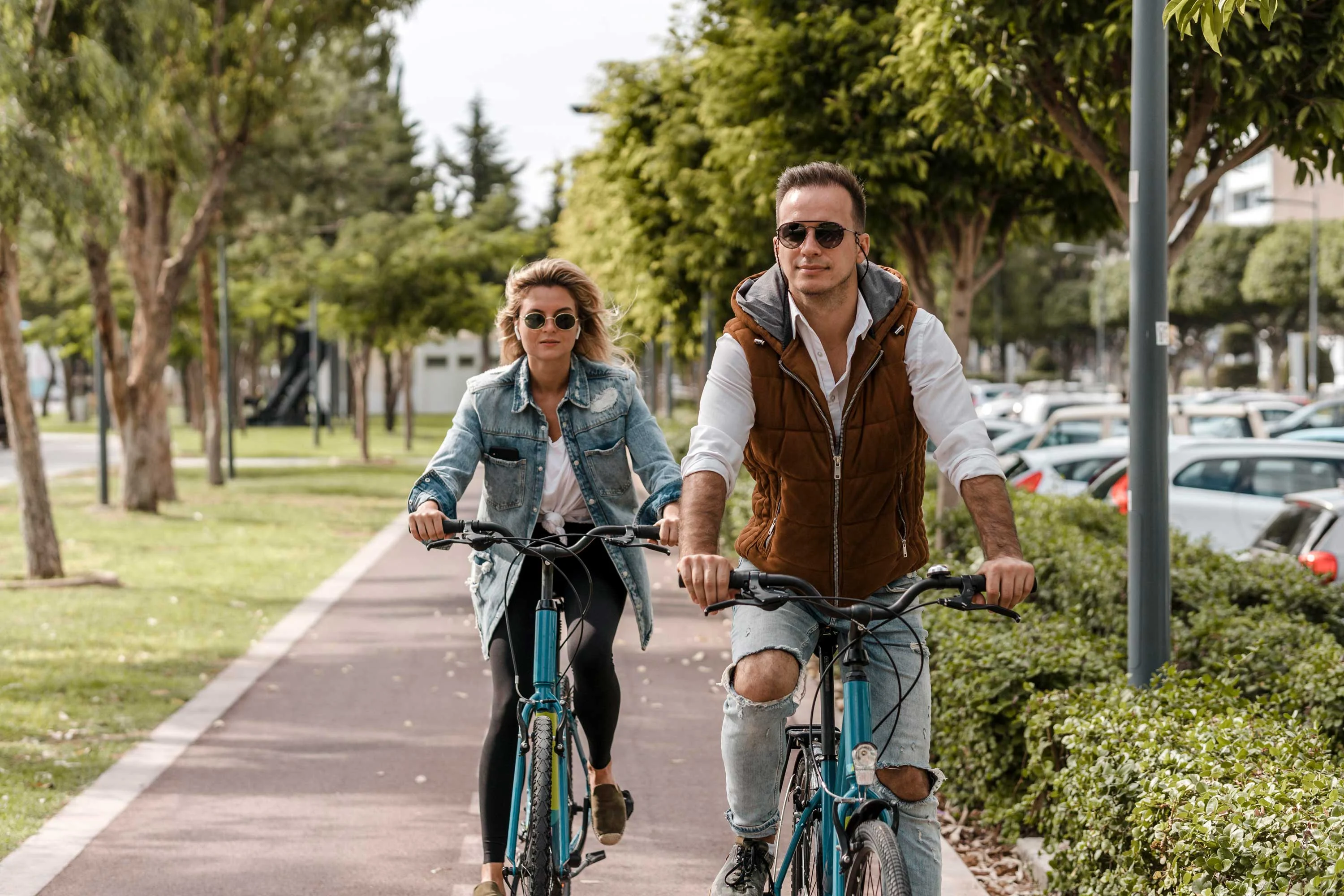 A couple riding bicycles along a path in a park, surrounded by trees and greenery. The man is wearing sunglasses and a casual outfit with a vest. The woman is also in sunglasses, dressed in a denim jacket, biking alongside him.