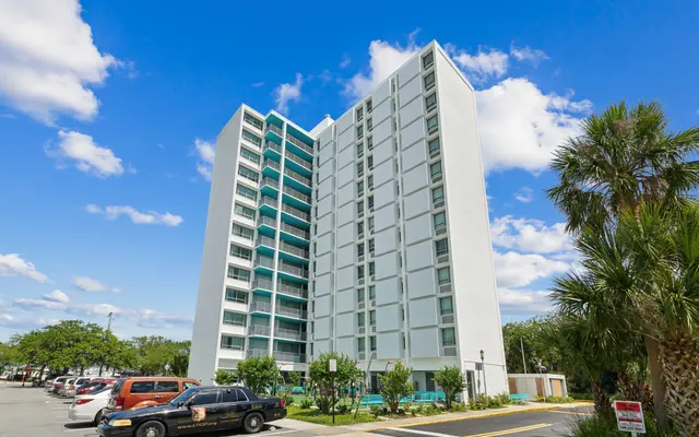 A tall, modern apartment building with a white façade, multiple balconies, and large windows, surrounded by palm trees and parked cars under a blue sky.