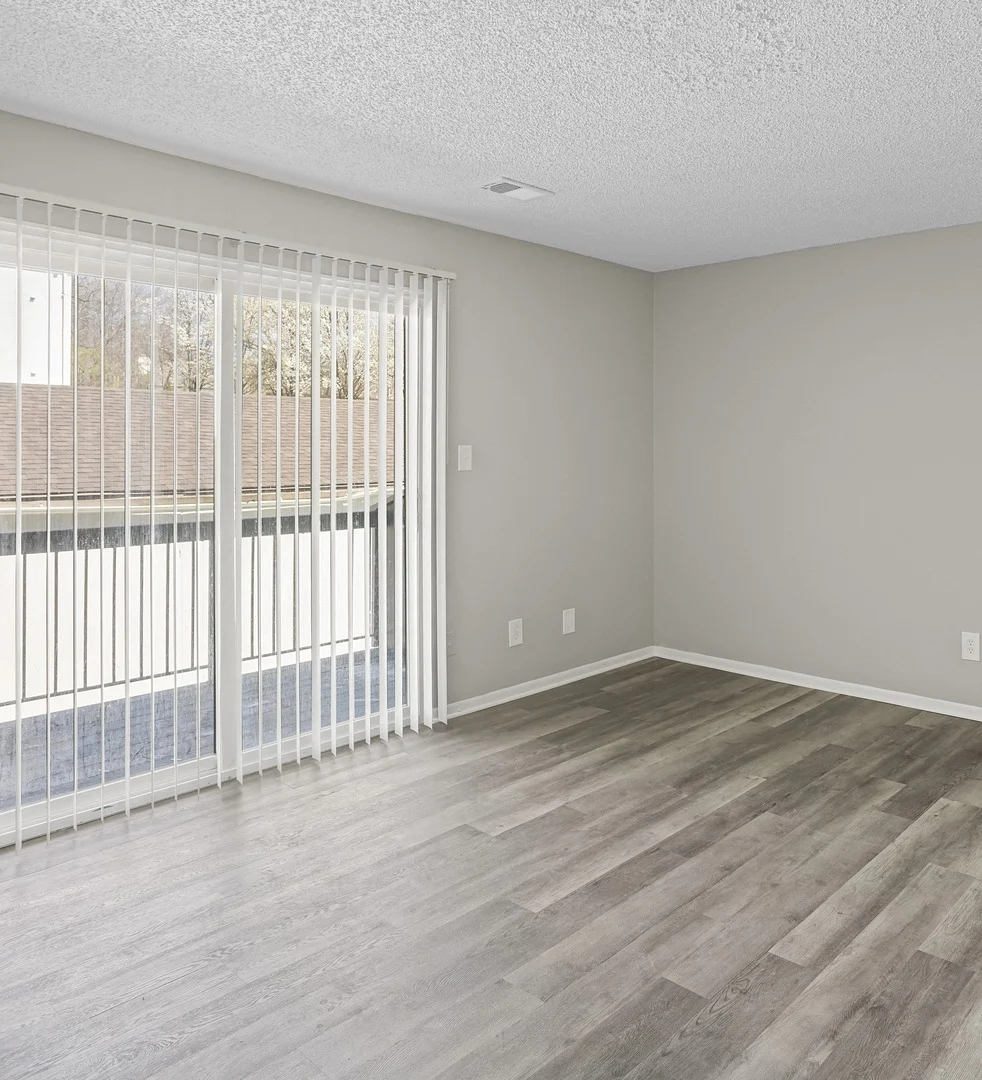 An empty living room with light gray walls and wood-like flooring. Large sliding glass doors with vertical blinds lead to a balcony.