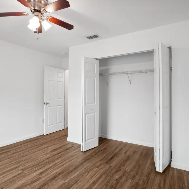 Empty Bedroom Interior with Closet An empty bedroom featuring a light wood floor, white walls, and a ceiling fan with wooden blades. There's a closet on the right with double doors opened, and a door on the left leading to another room.
