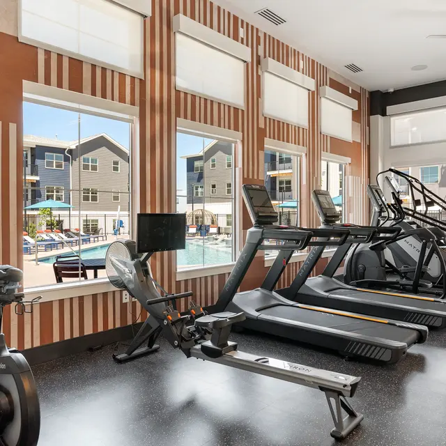A modern gym with exercise equipment including elliptical machines, stationary bikes, and a rowing machine, featuring large windows overlooking a pool area.