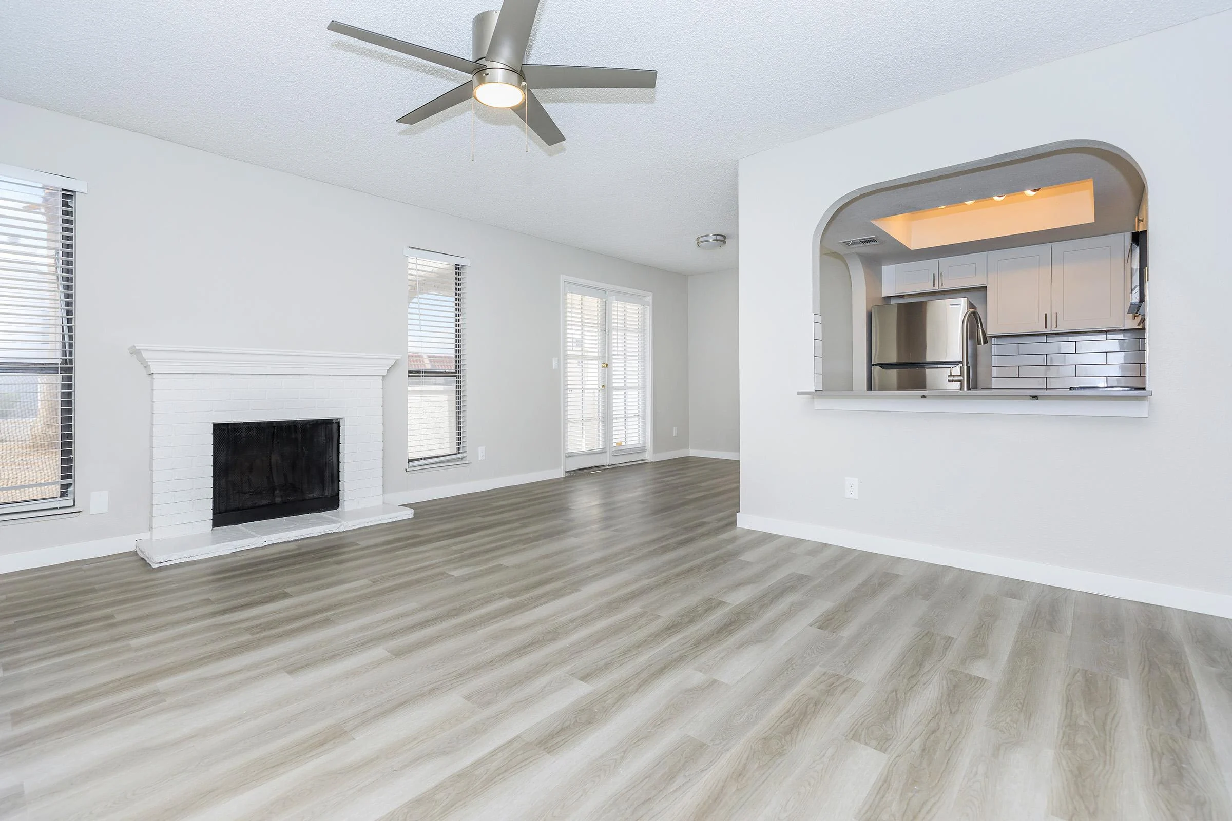 Bright, spacious living room with wooden floors, white walls, and a ceiling fan. Features a fireplace and a view into a kitchen area.