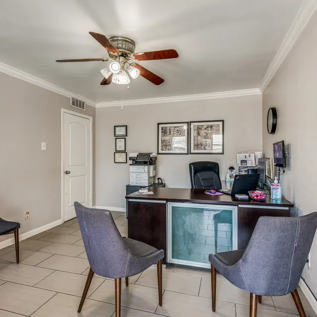 Modern Office Reception Area A modern office reception area with two gray chairs in front of a desk. The desk has a glass front and is equipped with a computer and office supplies. A wall with decorative art and a round mirror is visible, and a ceiling fan is overhead.