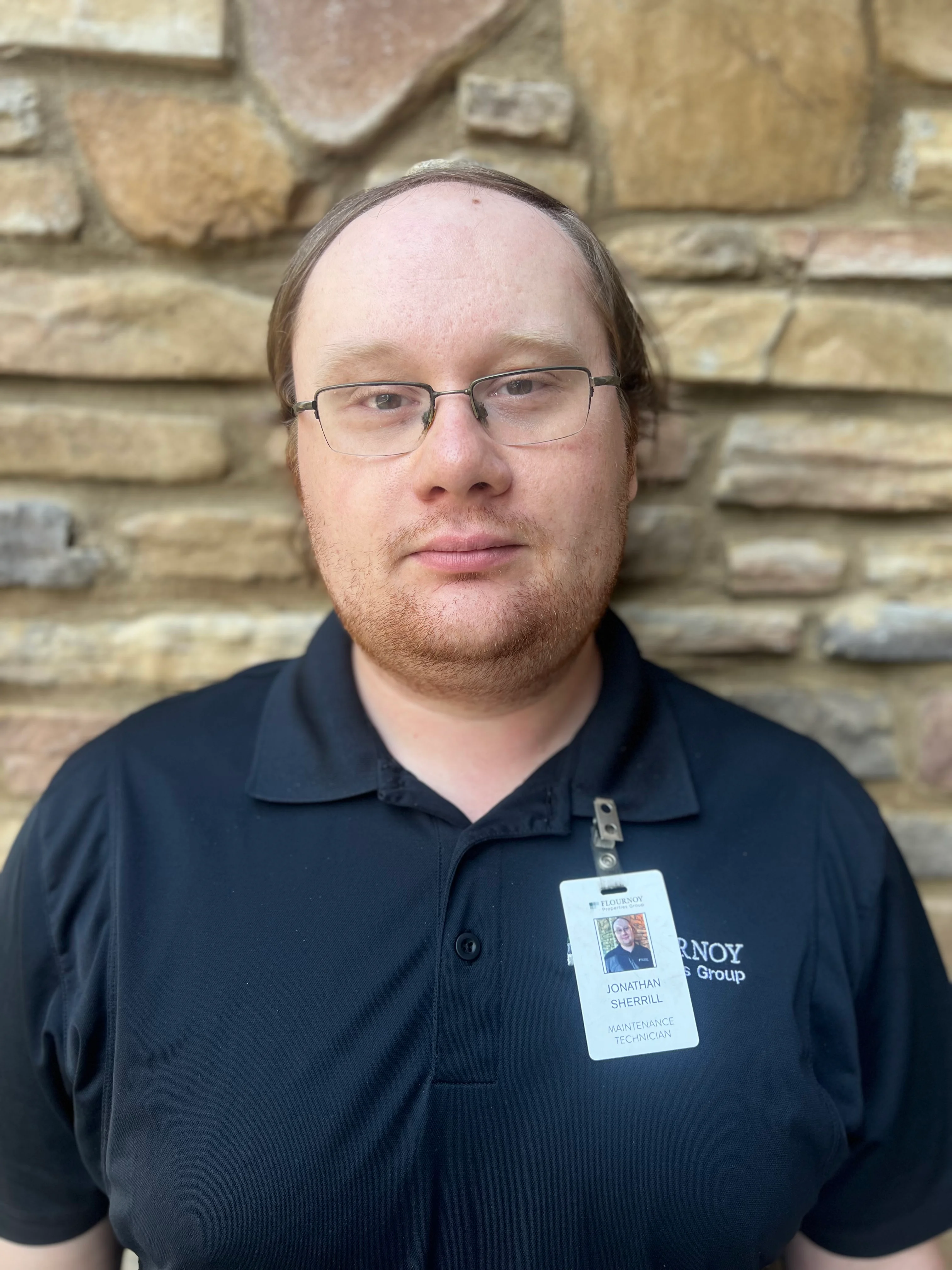 A man with glasses wearing a black polo shirt standing in front of a stone wall, sporting a badge attached to his shirt