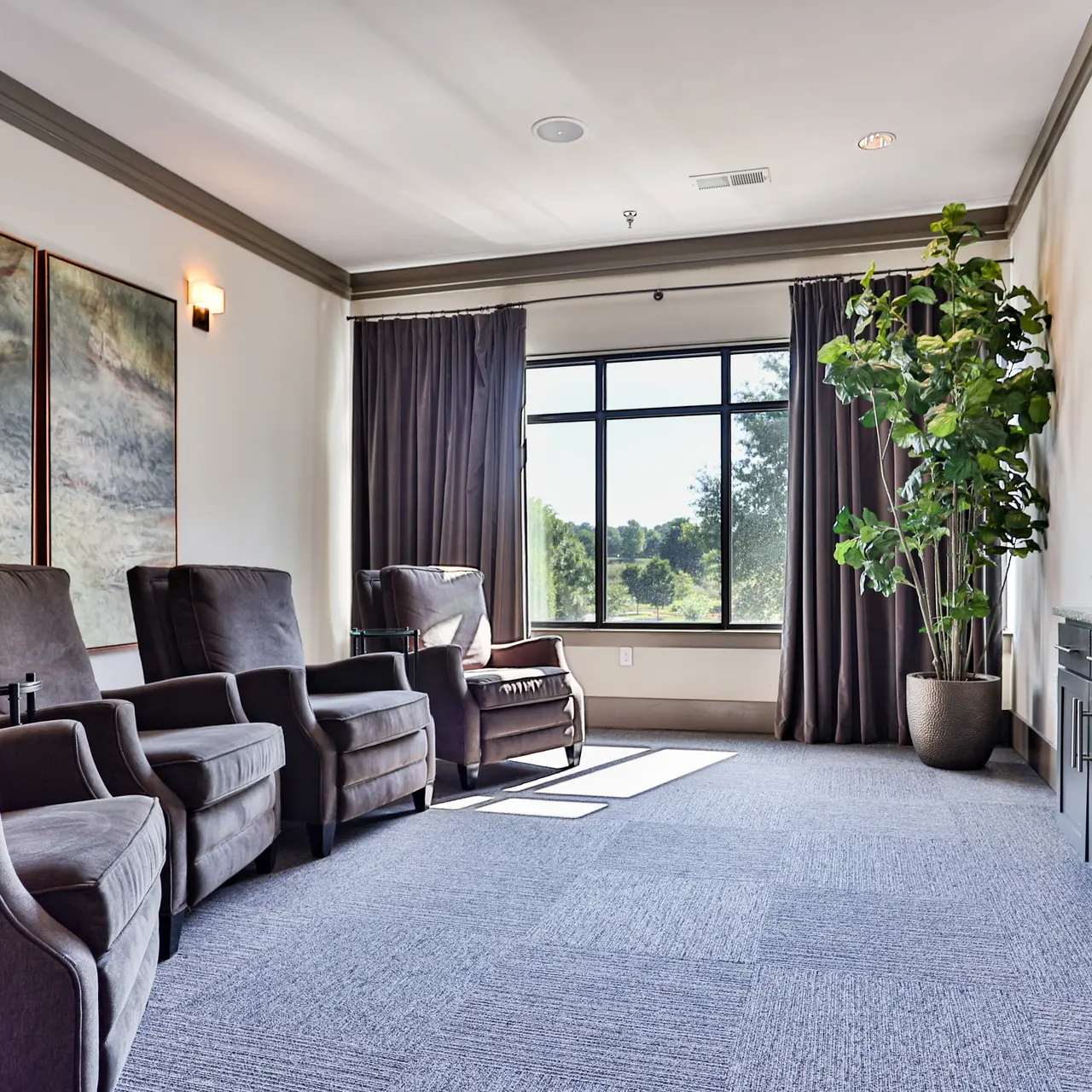 Modern Lounge Area A modern lounge area featuring two armchairs and a sofa beside large windows with a view of greenery. Artwork hangs on the walls, and there is a potted plant in the corner.