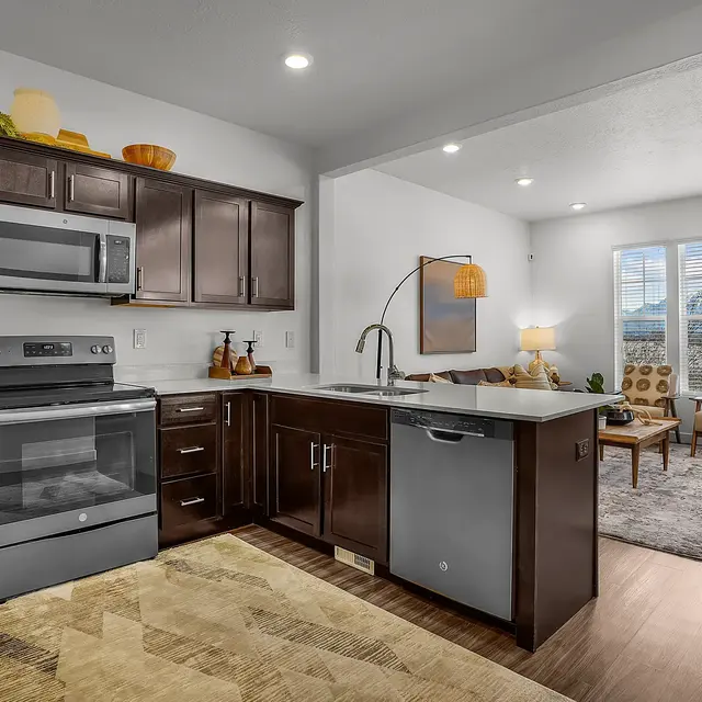 A modern kitchen with dark wooden cabinets, stainless steel appliances, and a minimalist design, adjacent to a cozy living room area featuring large windows and comfortable seating.