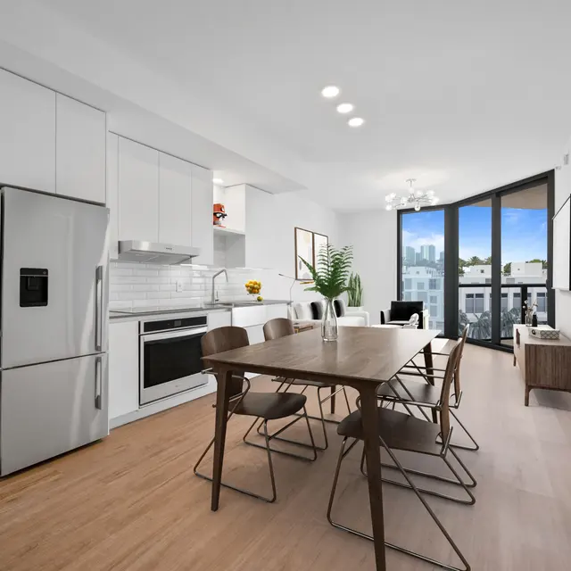 A modern kitchen and dining area featuring sleek white cabinetry, a refrigerator, an oven, and a dining table with metal chairs. Large windows provide natural light and a view of the outdoors.