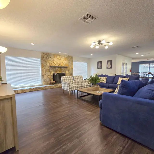 A spacious living room with a stone fireplace, blue sofas, and wooden flooring. Bright windows allow natural light to fill the area, while a dining space is visible in the background.