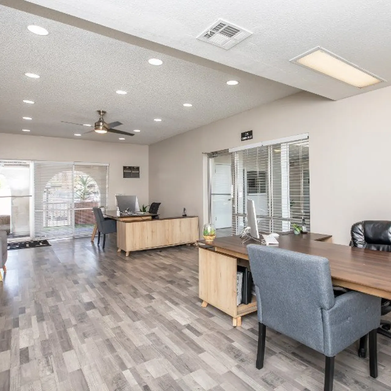 A modern office interior featuring a spacious layout with wooden flooring, two desk areas, and natural light coming from large windows. There are gray chairs and a fan on the ceiling.