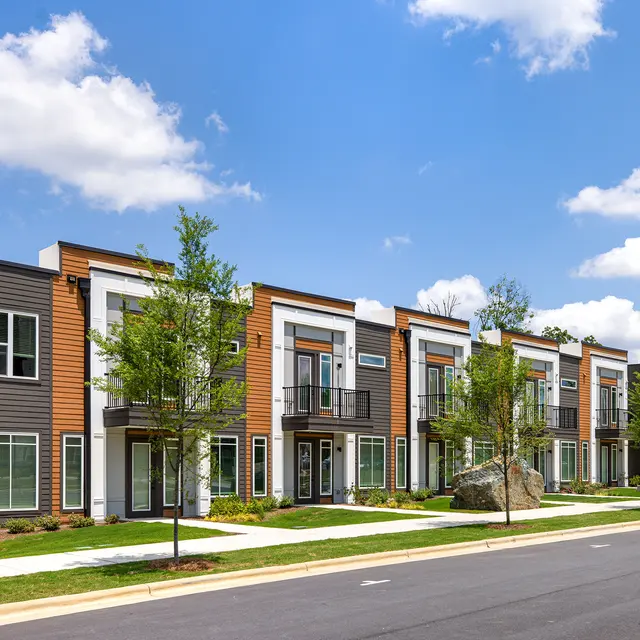 A row of modern multifamily townhouses with a mixture of gray and wooden exteriors, featuring balconies and surrounded by greenery under a clear blue sky.
