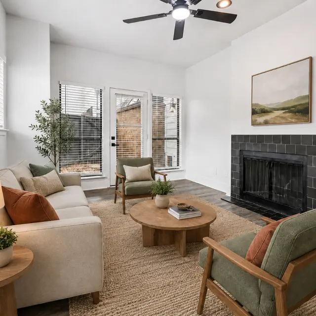 A cozy modern living room featuring a light-colored sofa, two armchairs, a round coffee table, and a fireplace. Large windows provide natural light, and a plant adds a touch of greenery.