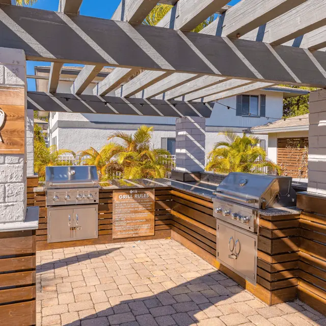 Inviting Outdoor Grilling Space An outdoor grilling area featuring two stainless steel grills under a wooden pergola, surrounded by landscaped greenery.
