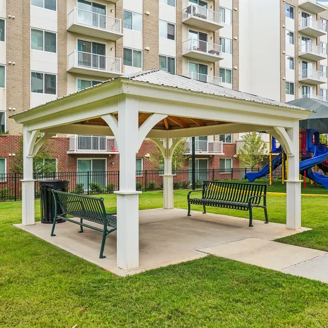 A shaded pavilion with benches in a grassy area, surrounded by trees and a playground in the background.