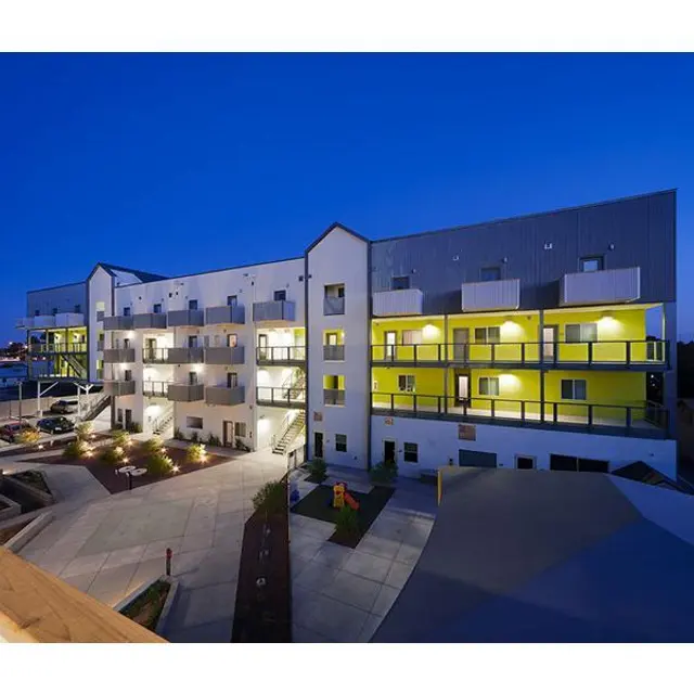 A modern multi-story apartment complex featuring yellow and white exterior with balconies and landscaped areas, captured during dusk with lights on.