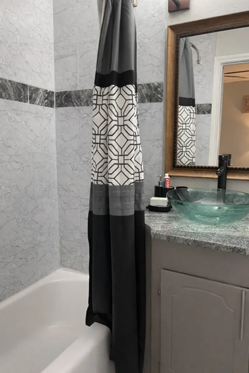 A bathroom featuring a tub with a shower curtain that has a geometric design. The sink area has a glass vessel sink and a modern faucet, with a large mirror and decorative elements in the background.