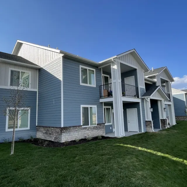 A multi-unit residential building with a light blue exterior and stone accents. The building has multiple balconies and windows, surrounded by well-maintained grass and a clear blue sky.