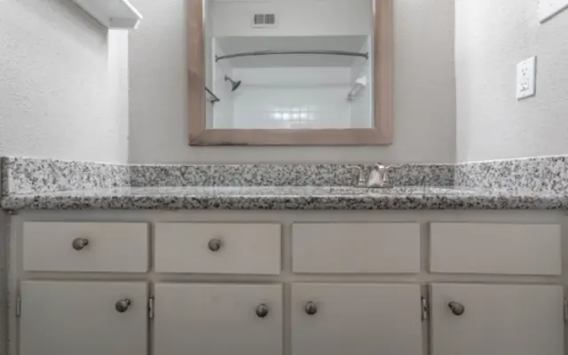 Modern Bathroom Vanity A bathroom counter with a granite surface, featuring a mounted mirror above and a faucet. There are several drawers below the counter, and the walls are light-colored.