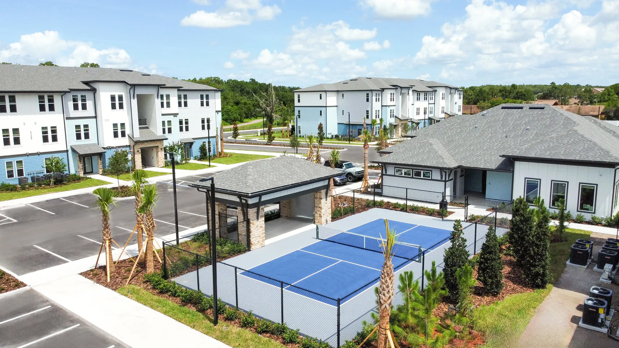 Aerial view of a modern apartment complex with blue tennis courts, landscaped gardens, and parking areas in the foreground.